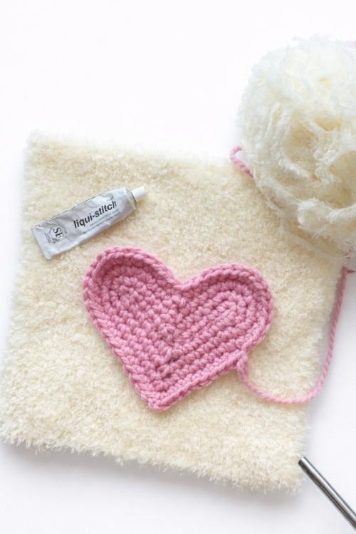 A pink crocheted heart with loose yarn rests on cream cloth next to a tube of glue labeled "liqui-stitch" and a cream yarn ball. Free crochet pattern