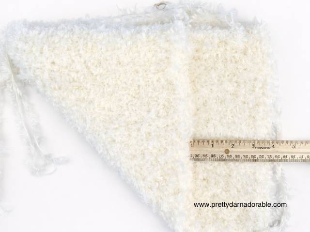 Close-up of a white, fluffy, triangular fabric piece with a ruler showing its length; attached string and website text in the corner. Free crochet pattern