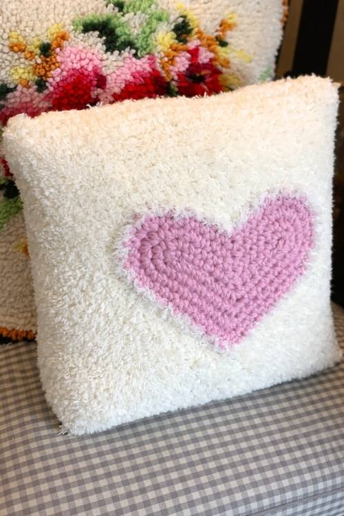 White crochet heart pillow with a pink heart on a gray and white checkered surface, colorful floral pillow in the background. No visible text. Free crochet pattern
