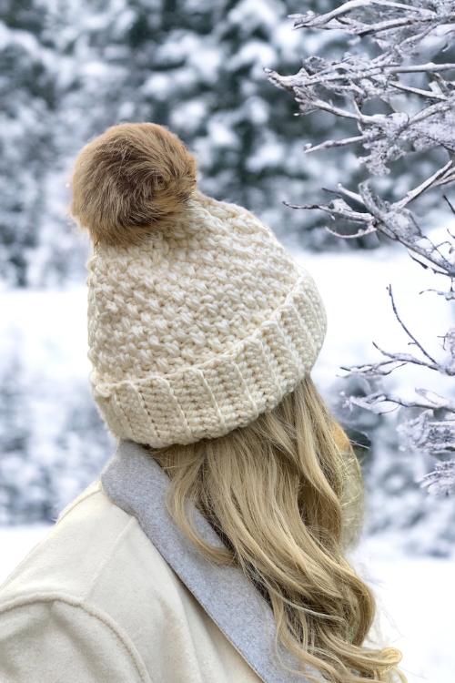 A person with long blonde hair and a cream hat with a brown pom-pom stands outside among snowy trees, facing away. Free crochet pattern
