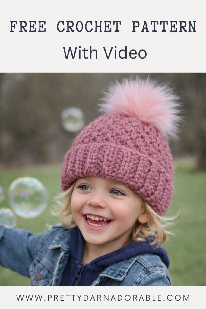 Smiling child outdoors in a pink crochet hat with pom-pom, bubbles floating. Text: Free Crochet Pattern With Video. www.prettydarnadorable.com. Free crochet pattern