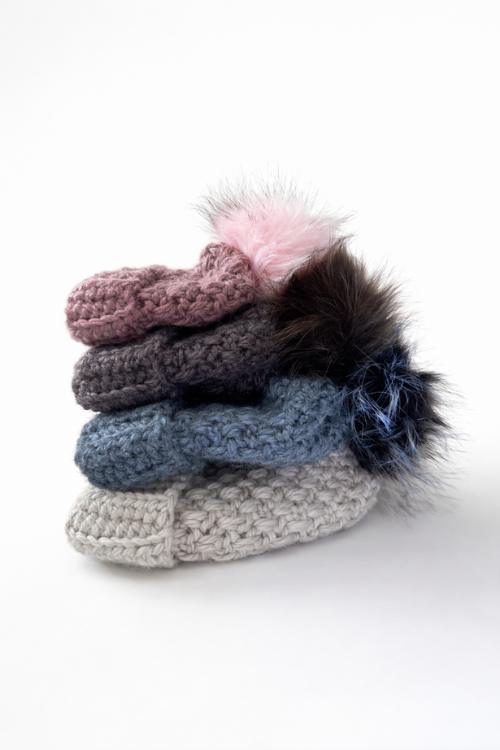 Four soft mittens in pink, gray, blue, and white with fur pom-poms at the wrists are stacked on a plain white background. Free crochet pattern