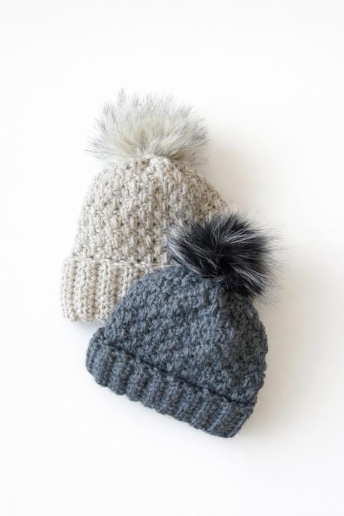 Two winter hats with furry pom-poms, one beige and one dark gray, overlap on a plain white background. No visible text. Free crochet pattern