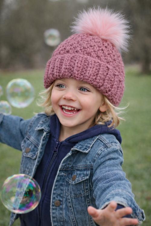 A smiling child in a pink hat with a pom-pom and denim jacket reaches for bubbles outside on grass. No visible text. Free crochet pattern