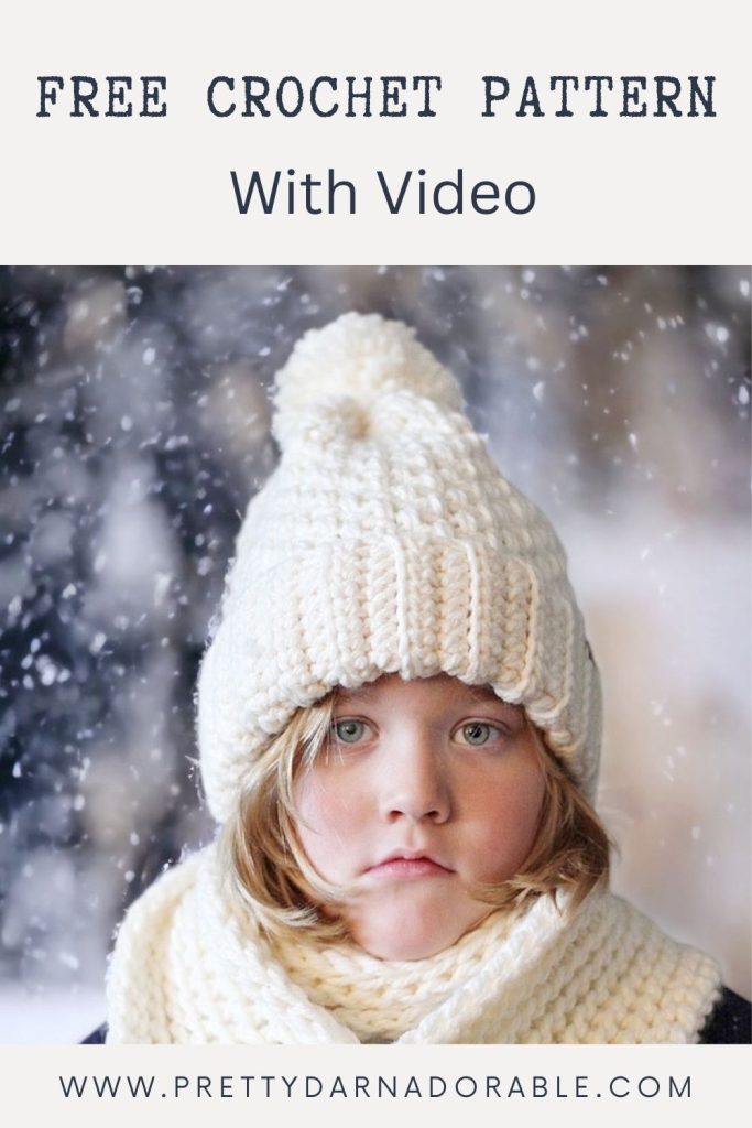 A young child in a thick white crochet hat and scarf stands outside in falling snow. Text: "Free Crochet Pattern With Video." www.prettydarnadorable.com. Free crochet pattern