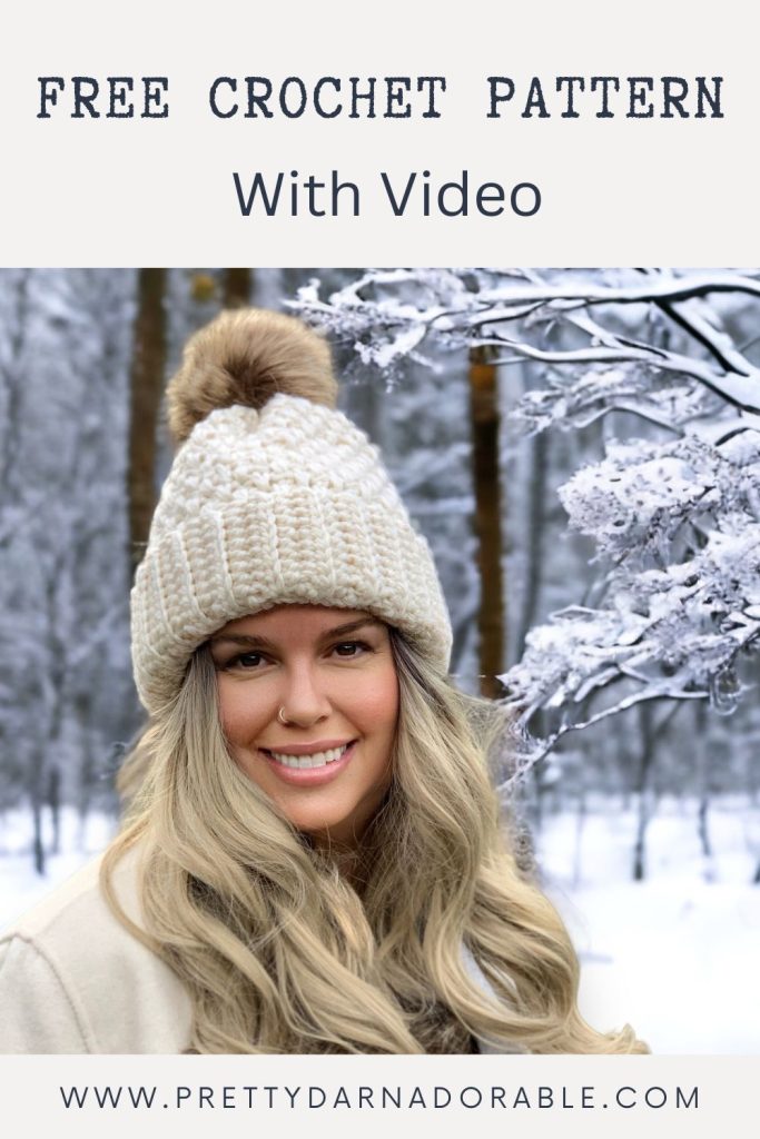 Smiling woman in a chunky white crochet hat with fur pom-pom, outdoors in snowy woods. Text: FREE CROCHET PATTERN With Video and www.prettydarnadorable.com. Free crochet pattern