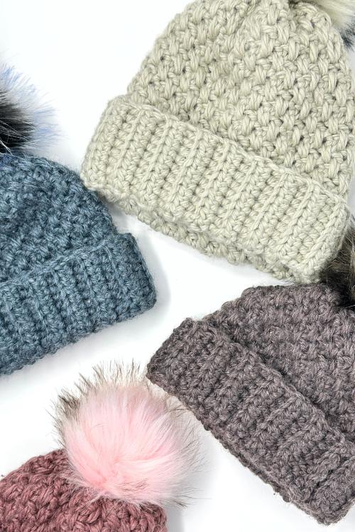 Four winter hats with ribbed brims and big furry pom-poms in cream, blue, brown, and mauve on a white background. Free crochet pattern