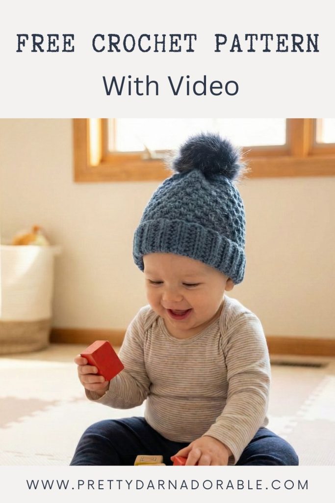A smiling baby in a blue crocheted pom-pom hat sits on a rug with red blocks; text reads “Free Crochet Pattern With Video,” www.prettydarnadorable.com. Free crochet pattern