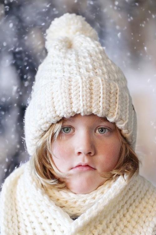 A young child with light hair in a white textured hat and scarf stands outside as snow falls, gazing seriously at the camera. Free crochet pattern