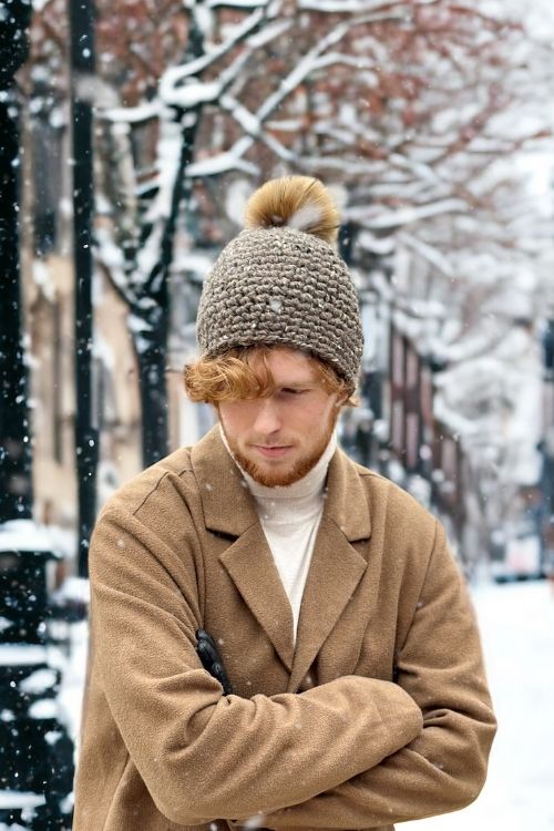 A young man with red hair and a beard stands in falling snow, wearing a brown coat and pom-pom hat; snowy trees and buildings behind. Free crochet pattern