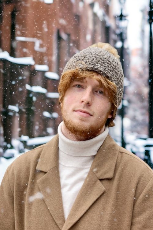 A young man with a light brown beard and wavy hair wears a beanie, beige coat, and white turtleneck on a snowy city street. Free crochet pattern