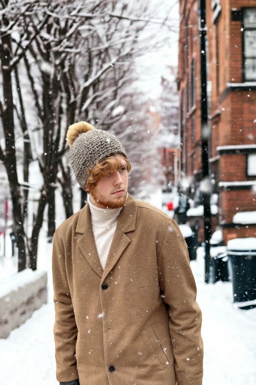 Young man in a tan coat and beanie walks on a snowy city street with bare trees and brick buildings; snowflakes fall around him. Free crochet pattern
