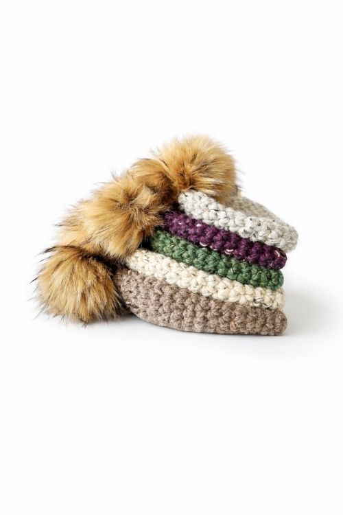Four stacked hats in beige, green, purple, and tan, each with a large brown faux fur pom-pom, set against a white background. Free crochet pattern