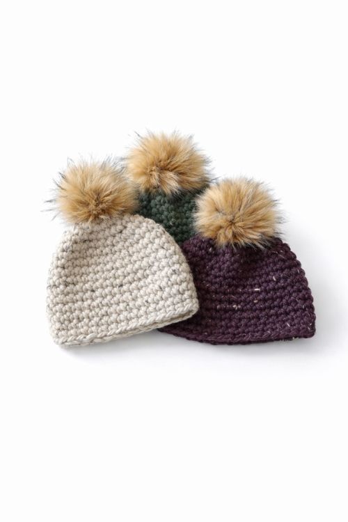 Three beanies in cream, green, and dark purple with faux fur pom-poms overlap on a plain white background. No text visible. Free crochet pattern