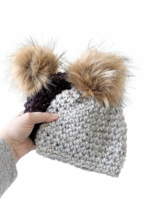 A hand holds a chunky hat with two large faux fur pom-poms, mostly light gray with a small section of dark gray or black yarn. Free crochet pattern