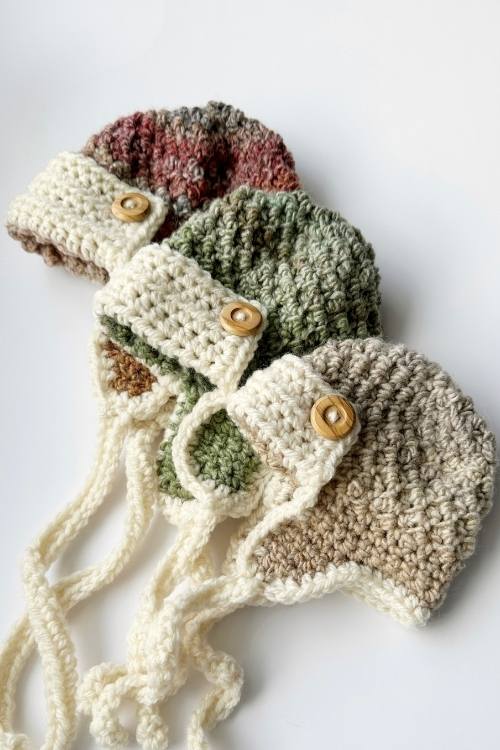 Three crochet aviator hats in green, beige, and maroon with wooden buttons and braided ties, shown on a white background. Free crochet pattern