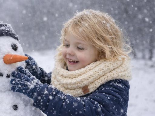 A smiling blonde child in a cream scarf touches a snowman with a carrot nose and black hat outside in falling snow. Free crochet pattern