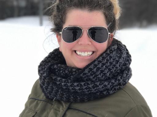 Woman smiling outside in snow, wearing aviator sunglasses, a dark scarf, and olive green jacket; snowy background is blurred. Free crochet pattern