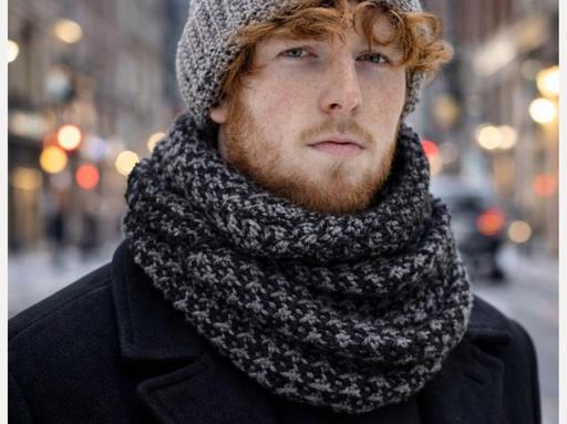 Young red-haired man with a beard in a gray hat, black-and-gray scarf, and dark coat stands on a city street at night; blurred lights and buildings in background. Free crochet pattern