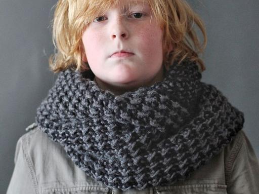 A blond-haired child in a thick, dark gray textured scarf and gray jacket stands against a plain gray background. Free crochet pattern