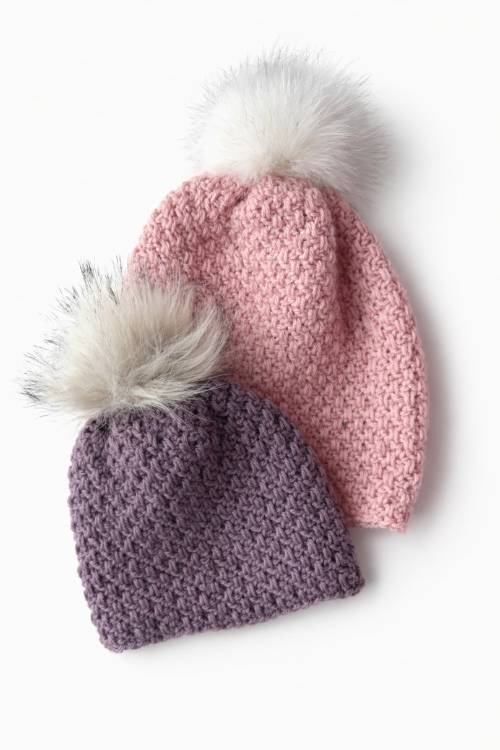 Two hats with chunky texture and white faux fur pom-poms, one pink and one lavender, on a white background. Free crochet pattern