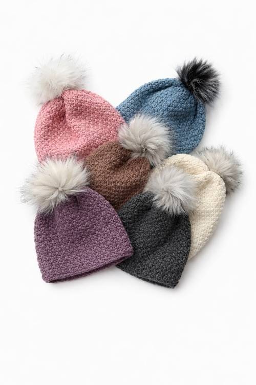 Five beanies with faux fur pom-poms in pink, blue, brown, cream, and purple are fanned out on a white background. Free crochet pattern