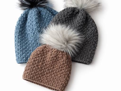 Three beanies in blue, dark gray, and brown with large faux fur pom-poms overlap on a white background. No text visible. Free crochet pattern