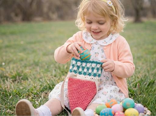 Smiling young girl on grass puts painted Easter egg in crochet carrot bag; floral dress, pink cardigan, colorful eggs nearby. Free crochet pattern