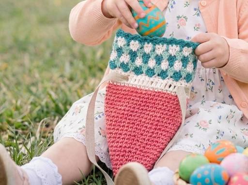 A child in a floral dress and pink sweater sits on grass, placing an Easter egg into a crocheted carrot bag; decorated eggs nearby. Free crochet pattern