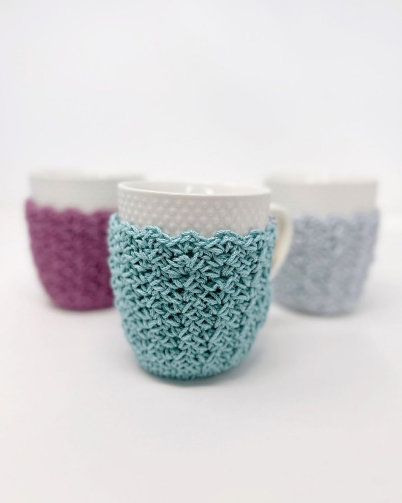 Three white mugs with blue, purple, and gray crochet cozies rest on a white surface, the blue cozy featured in front. Free crochet pattern