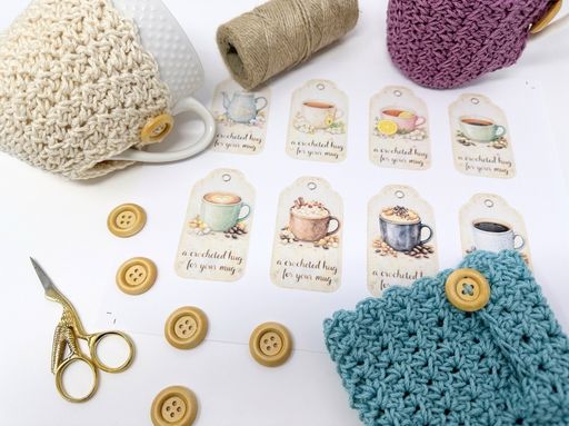 Colorful crocheted mug cozies with buttons, twine, tags reading &ldquo;a crocheted hug for your mug,&rdquo; and two mugs on a table. Free crochet pattern