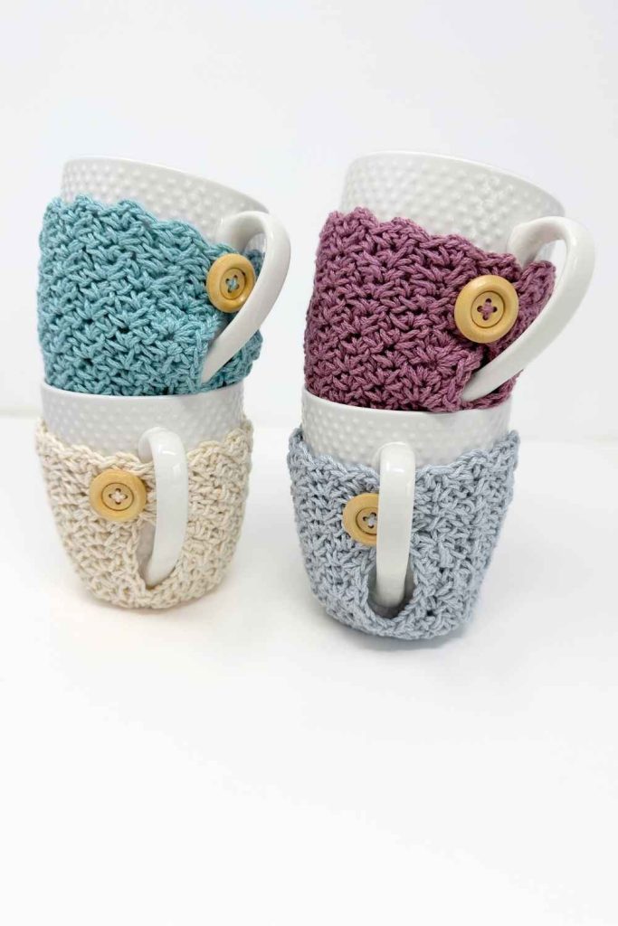 Four white mugs stacked in pairs, each in a beige, blue, or purple crochet cozy with a wooden button on a plain white background. Free crochet mug cozy pattern