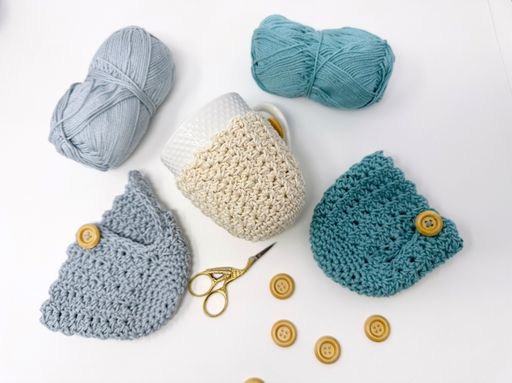 Two yarn balls, a mug in a crochet cozy, two buttoned pieces, scissors, and buttons on a white surface; no visible text. Free crochet pattern