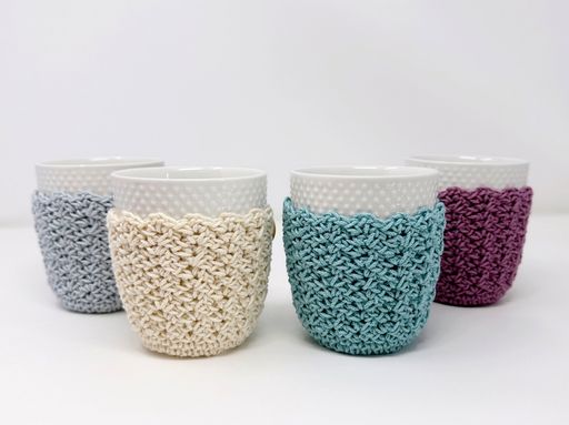Four white ceramic mugs with polka dots, each in a different colored crochet cozy&mdash;gray, cream, blue, and purple&mdash;on a white background. Promotional text: Try a free crochet mug cozy pattern to recreate this charming look at home. Free crochet pattern