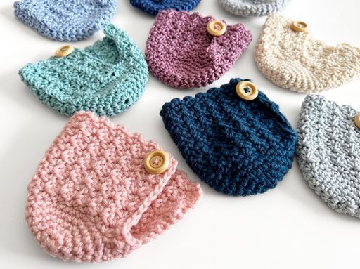Small crocheted pouches in pastel pink, blue, purple, and cream with wooden buttons on a white surface; &ldquo;Free crochet mug cozy pattern&rdquo; text visible. Free crochet pattern