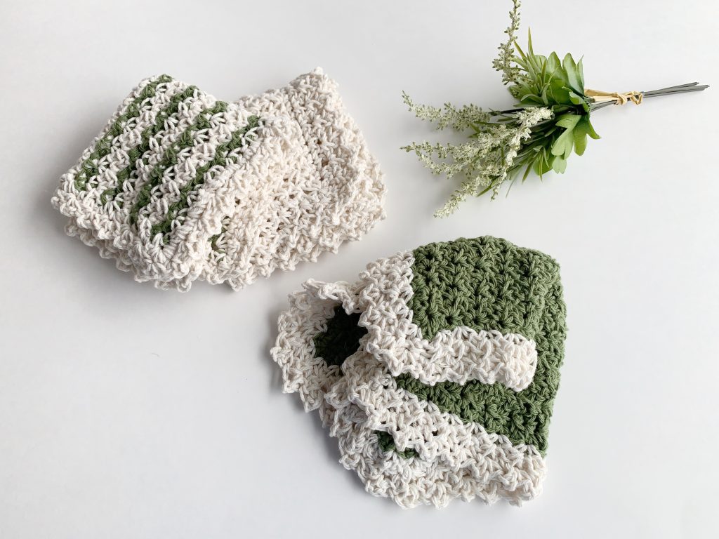 Two crocheted washcloths, one white and one green, rest on a white surface beside a small sprig of faux greenery and white flowers. Free crochet pattern