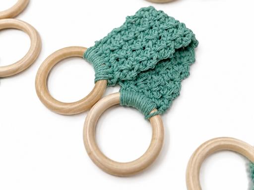 Two wooden rings linked by a wide teal macramé strap, with extra wooden rings on a plain white background. No text visible. Free crochet pattern