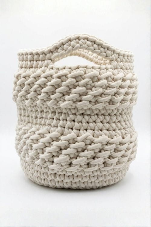 A cream-colored, handwoven basket made from thick, chunky yarn features a textured spiral pattern and an oval cutout handle near the top; visible text promotes a free crochet basket pattern for cozy crafting projects. Free crochet pattern
