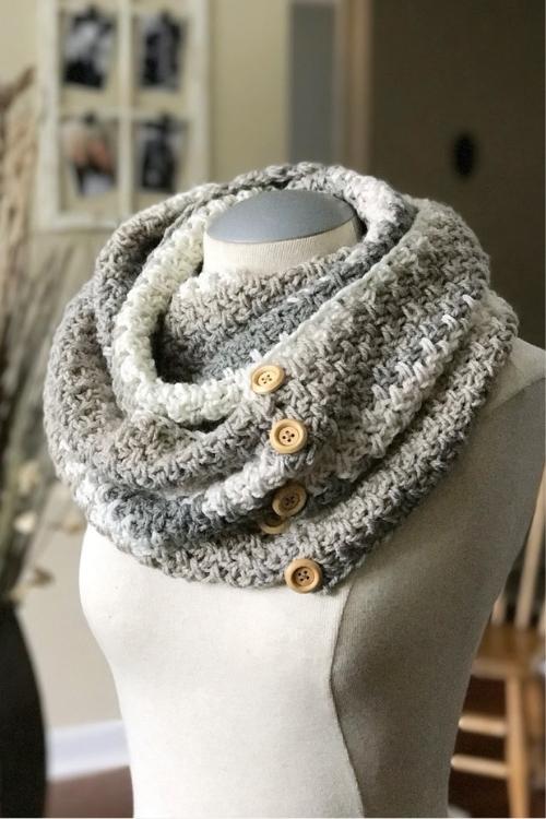 A chunky gray and white textured cowl scarf with three wooden buttons is shown on a dress form in a softly lit, cozy room. Free crochet pattern