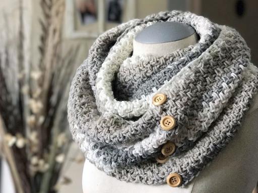 A gray and white textured cowl scarf with wooden buttons is displayed on a dress form, with a soft, neutral background featuring decor and dried plants. No visible text or promotional info appears in the image. Free crochet pattern