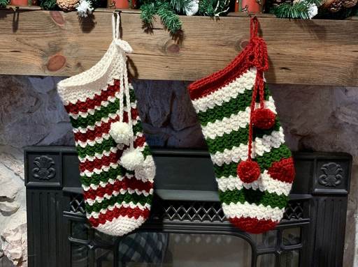 Two Christmas stockings with red, green, and white stripes and two pom-poms each hang from a wooden mantel above a fireplace. The stockings have a crocheted texture; no visible text or promotional info appears in the image. Free crochet pattern