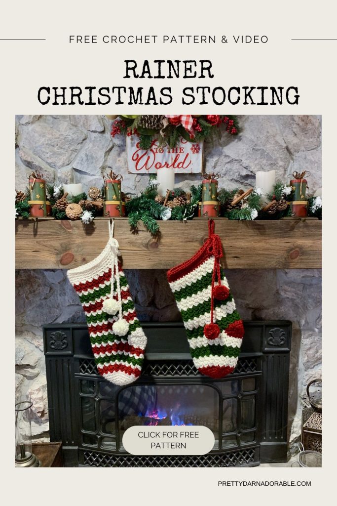 Two striped crochet Christmas stockings, one red and one green, hang from a mantel decorated with garlands, candles, and ornaments above a fireplace with a sign that reads "To the world. Free crochet pattern
