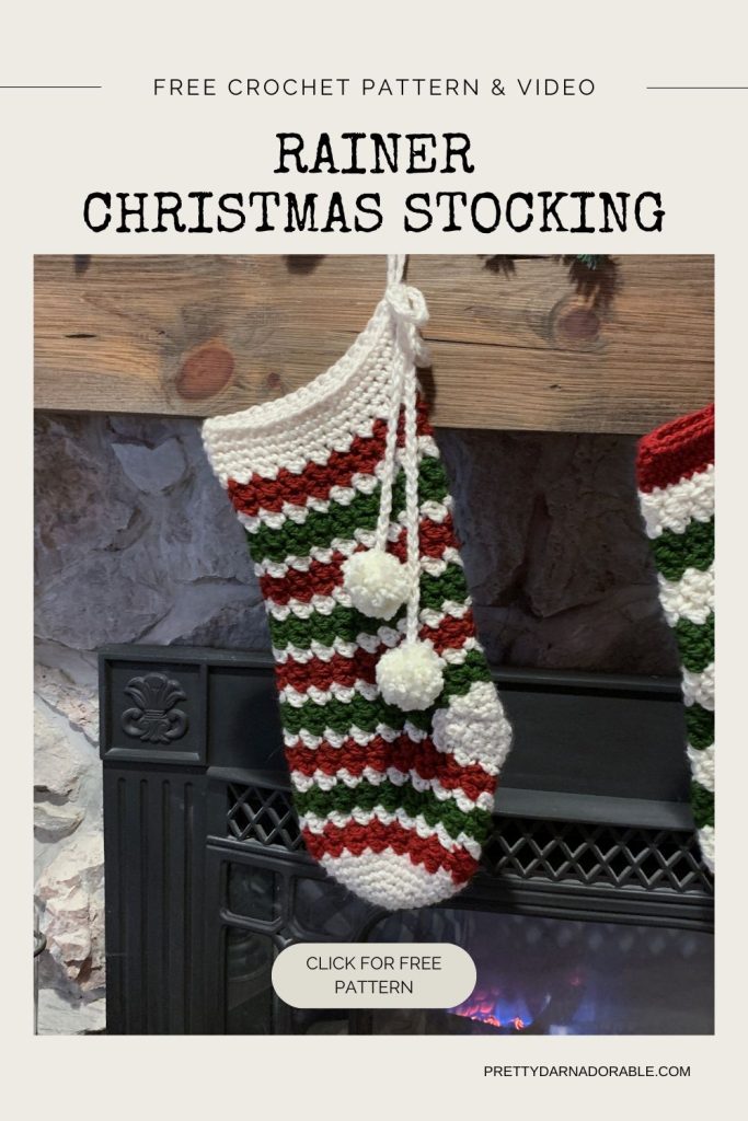 A handmade Christmas stocking with red, green, and white stripes and three yarn pom-poms hangs by a fireplace. Visible text says: Rainer Christmas Stocking, free crochet pattern & video. Free crochet pattern
