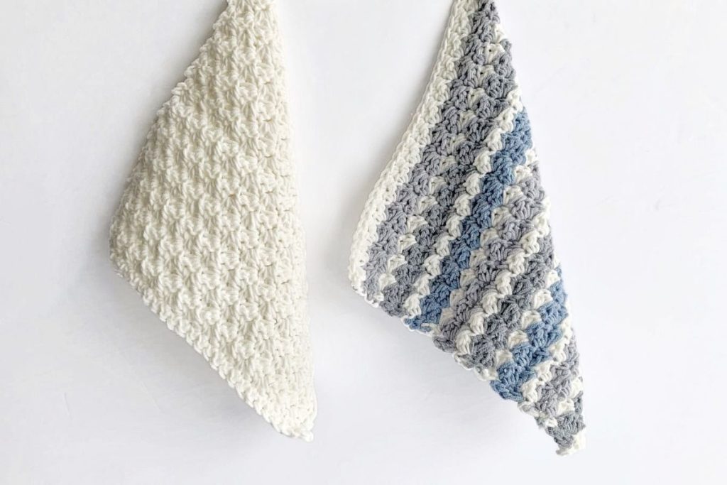 Two crocheted dishcloths with a bumpy, textured pattern hang on a white wall; one is solid off-white, and the other features stripes of white, grey, and blue. No visible text or promotional information appears in the image. Free crochet pattern