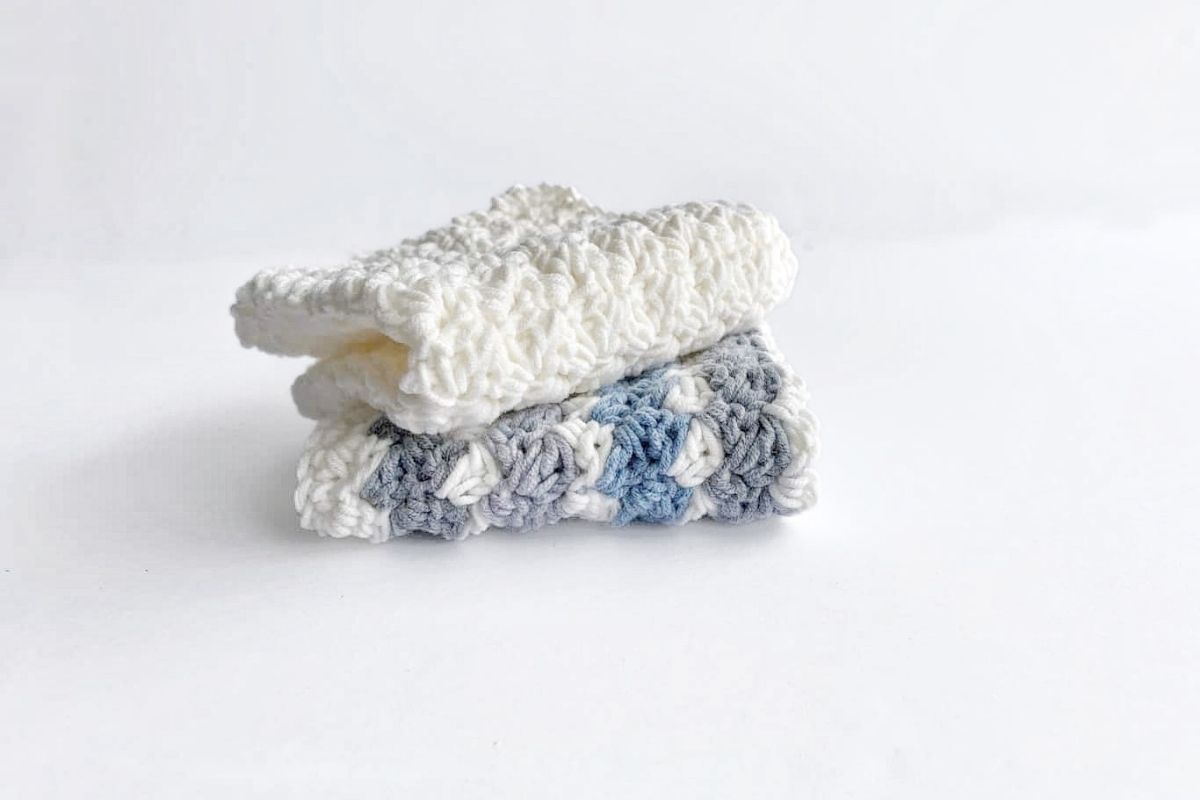 Two folded crocheted washcloths are stacked on a white surface; the top one is off-white, and the bottom has a pattern of white, gray, and blue yarn. No visible text or promotional information appears in the image. Free crochet pattern