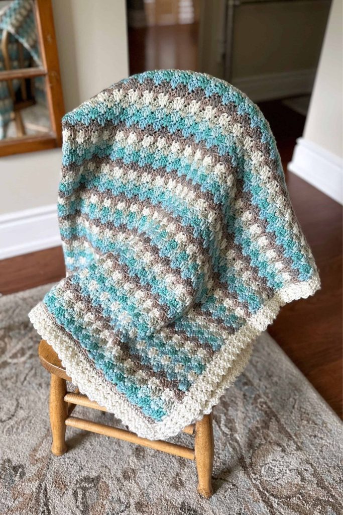 A striped crocheted blanket in blue, brown, and white is draped over a small wooden chair placed on a patterned rug in a softly lit, cozy room. No visible text or promotional information. Free crochet pattern