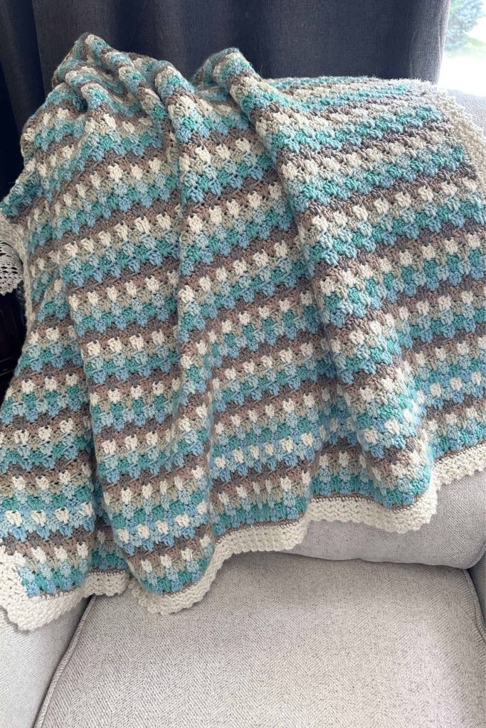 A striped crocheted blanket in blue, gray, brown, and white with a scalloped white edge and textured pattern is draped over a light gray armchair; dark curtains and a window are in the background. No visible text or promotional info. Free crochet pattern