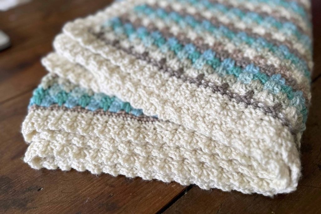 Folded crocheted blanket with cream, teal, gray, and brown stripes on a wooden surface; no visible text or promotional details. Free crochet pattern
