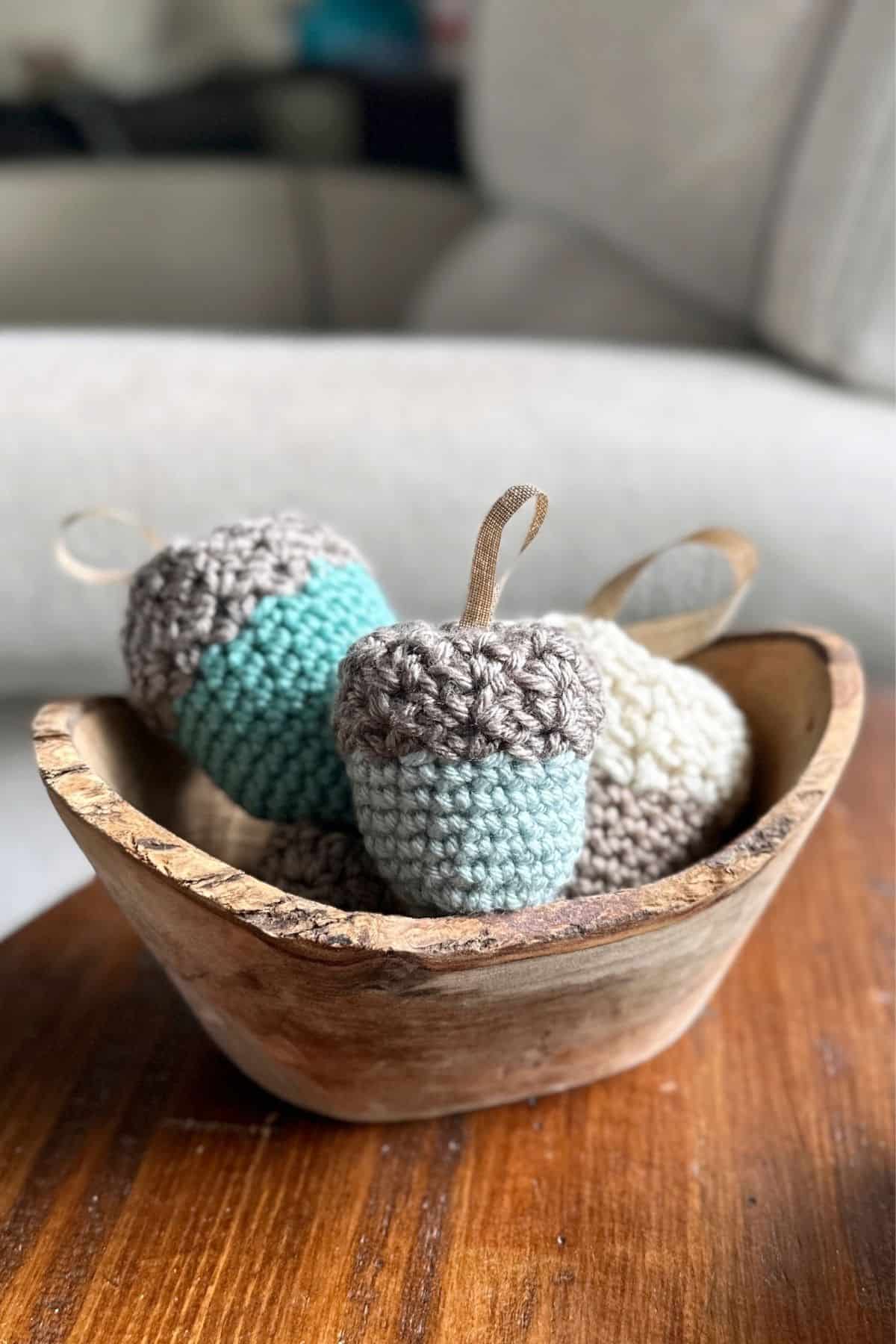 A wooden bowl displays three crocheted acorns in blue, gray, and cream, each with a loop for hanging; the background features a blurred couch. Text on the image promotes these as beginner-friendly crochet projects or ideas for trying out a free crochet pattern. Free crochet pattern