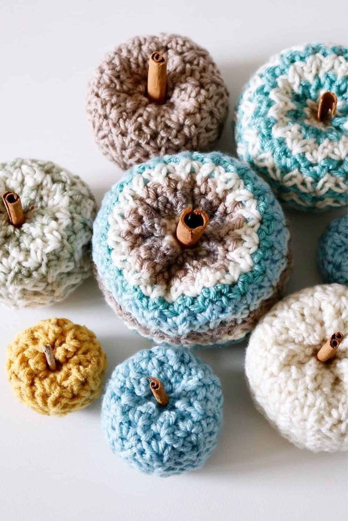 Pastel-colored crocheted pumpkins with cinnamon stick stems are arranged on a white surface, displaying the Cedar Ridge Pumpkins free crochet pattern. Free crochet pattern